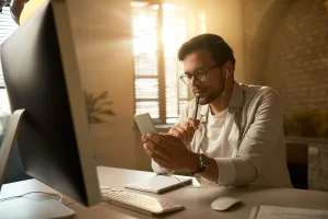 Empresário pensativo mandando mensagens de texto no celular enquanto trabalhava em um computador em sua mesa de escritório