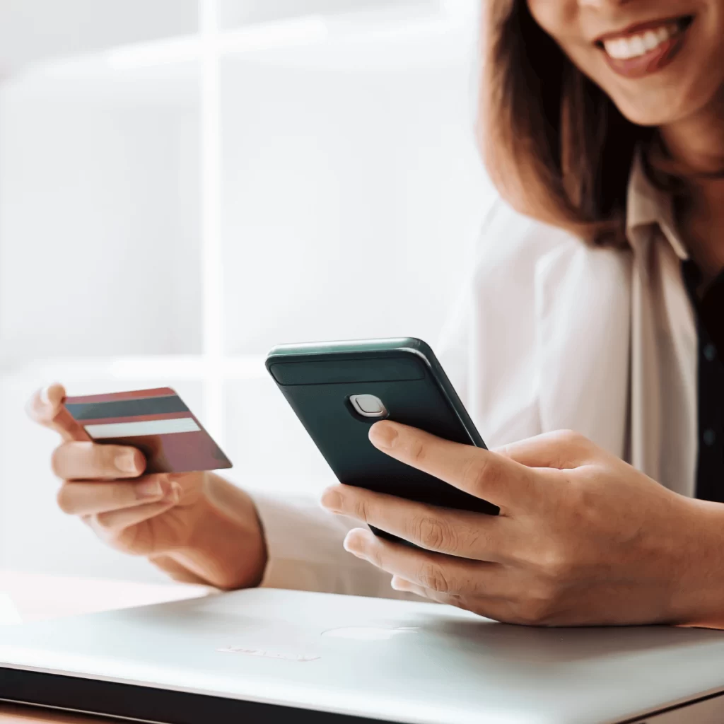 Mulher sorridente realizando um pagamento online com cartão de crédito e smartphone, destacando a facilidade e conveniência das transações digitais. No fundo, um laptop aberto sugere um ambiente de trabalho moderno.