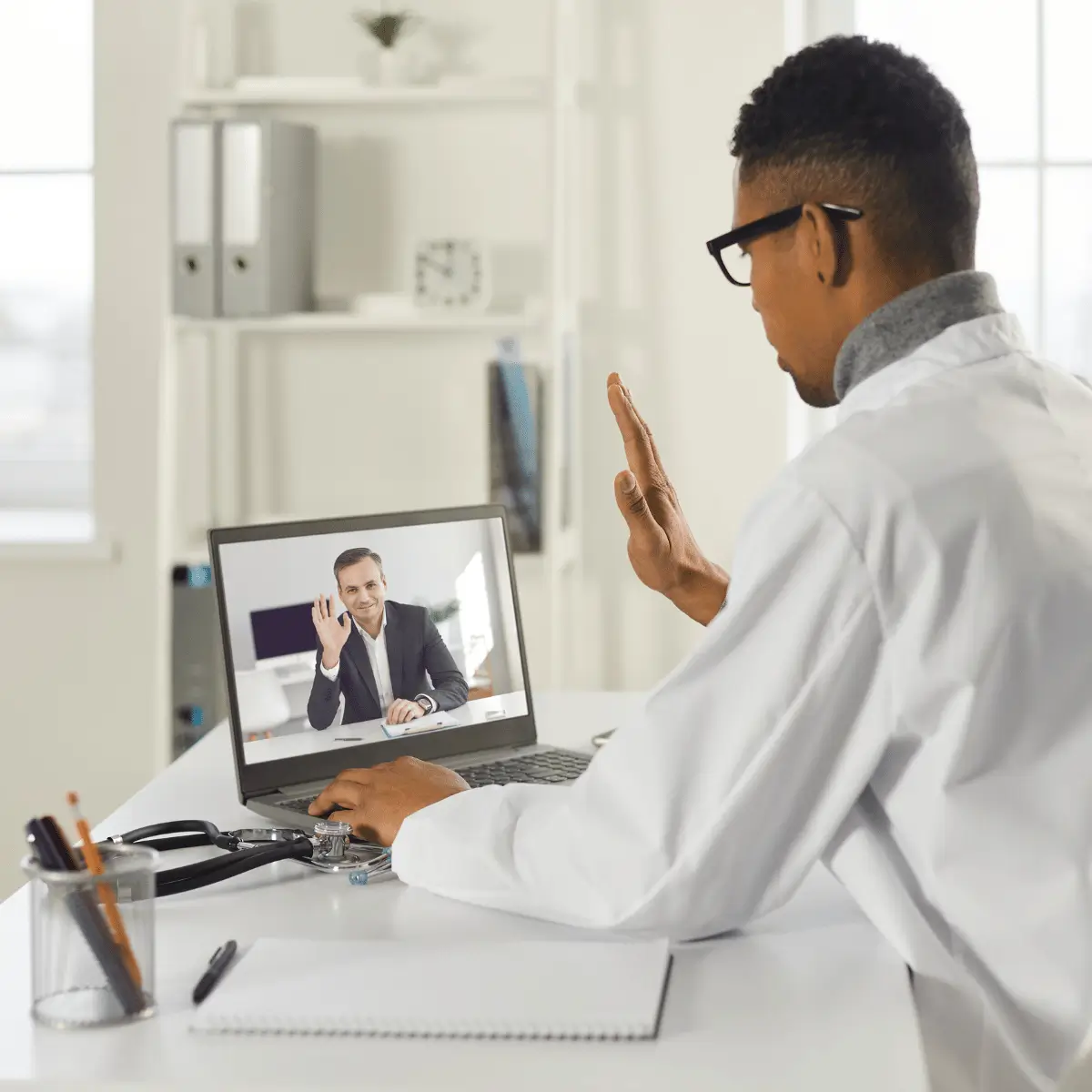 A imagem mostra um profissional da saúde, provavelmente um médico, usando um jaleco branco e óculos, participando de uma consulta virtual através de um laptop. Ele está levantando a mão em um gesto amigável de cumprimento, enquanto na tela do laptop aparece outro homem, bem-vestido, também acenando. A cena sugere uma consulta médica online ou uma reunião profissional na área da saúde, destacando a acessibilidade e a modernidade proporcionadas pela telemedicina. O ambiente é claro e organizado, com uma prateleira ao fundo e materiais de escritório sobre a mesa.